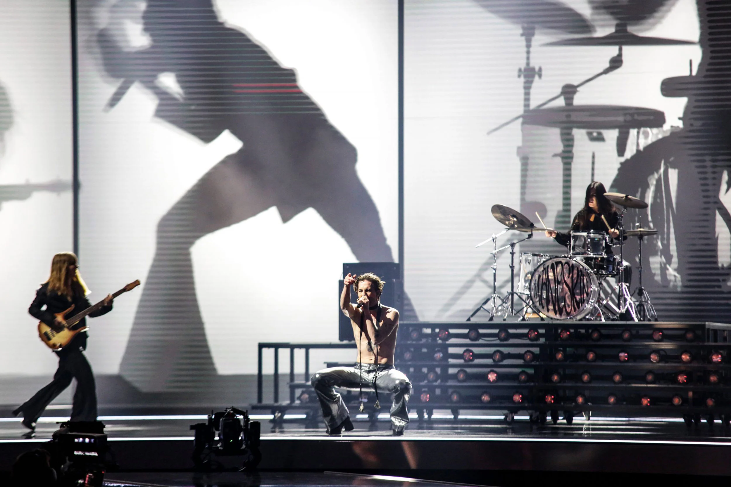 Singer crouching at the front of the stage, guitarist and drummer behind, band silhouettes projected on the backdrop – Måneskin at Eurovision 2021, Rotterdam, Netherlands – concert stage design