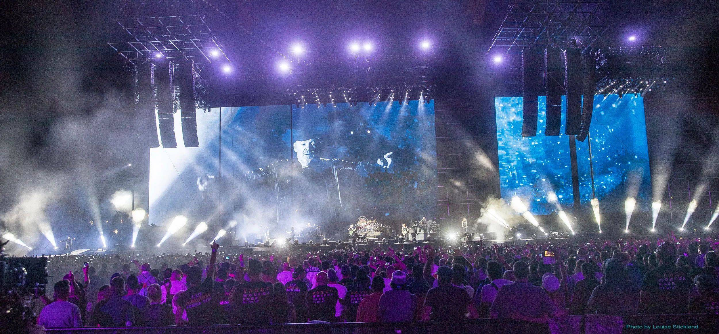 Singer filmed on the main screen and a blue-tinted side screen, white spotlights sweeping the audience – Vasco Rossi Modena Park, Italy, 2017 – concert stage design