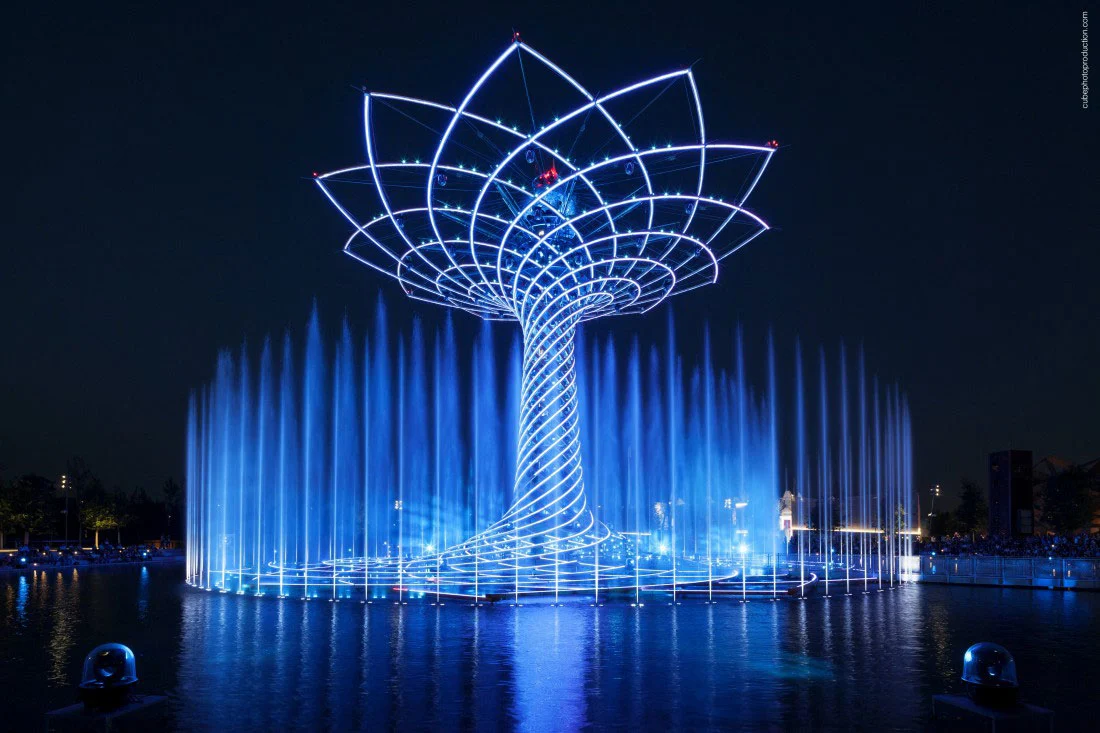 Tree of Life lit in blue at night, fountain jets rising around its base and reflected in the lake – Albero della Vita at Expo 2015, Milan, Italy – installation design