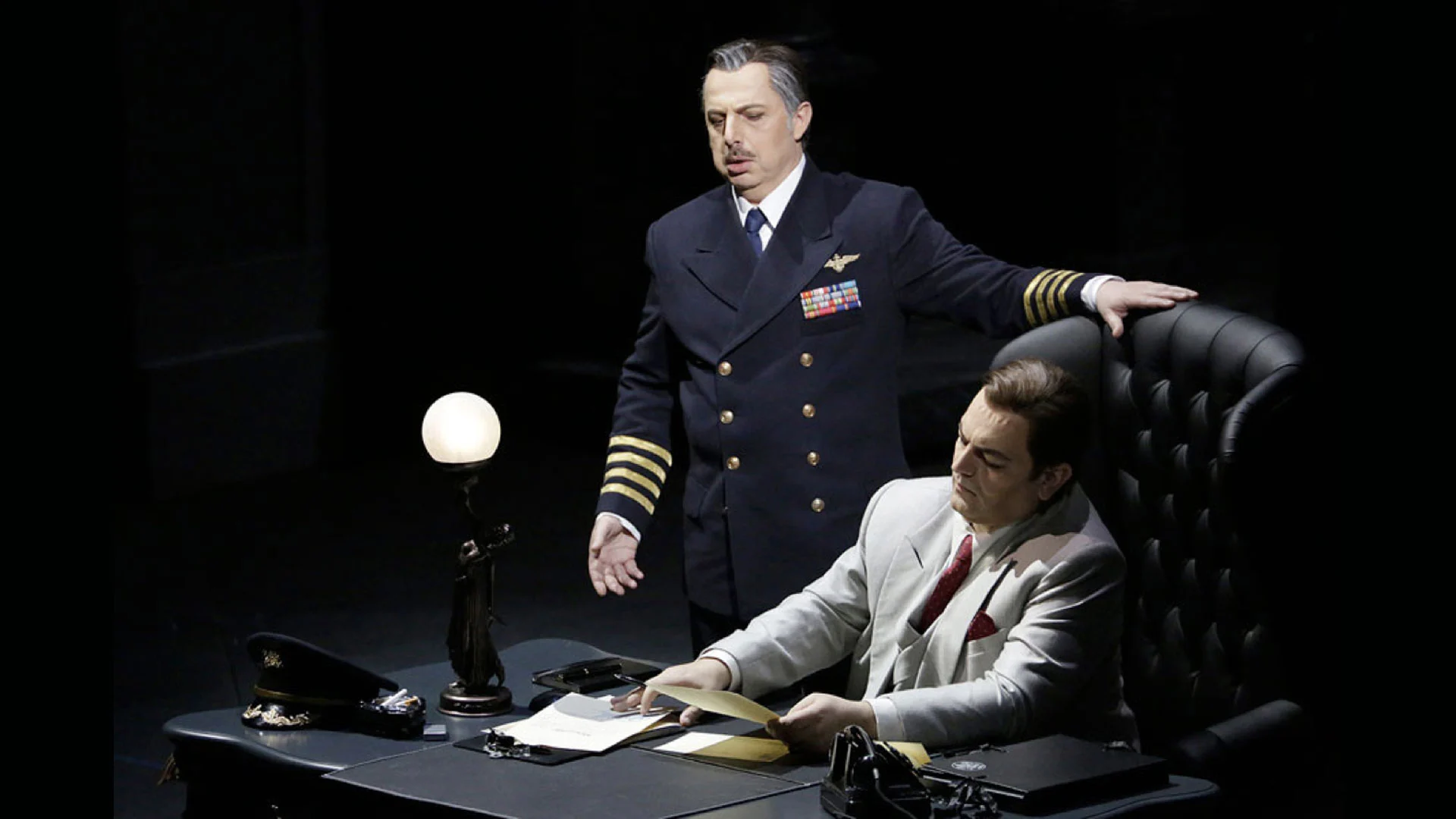 Officer in a navy uniform standing beside a seated man in a pale suit at a desk with a globe lamp – Un Ballo in Maschera at the Bolshoi Theatre, Moscow, Russia, 2018 – opera set design