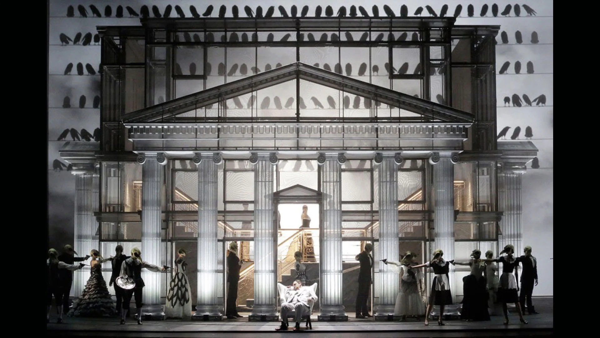 Neoclassical temple facade set with birds silhouetted on the walls, masked guests lined up on either side – Un Ballo in Maschera at the Bolshoi Theatre, Moscow, Russia, 2018 – opera set design