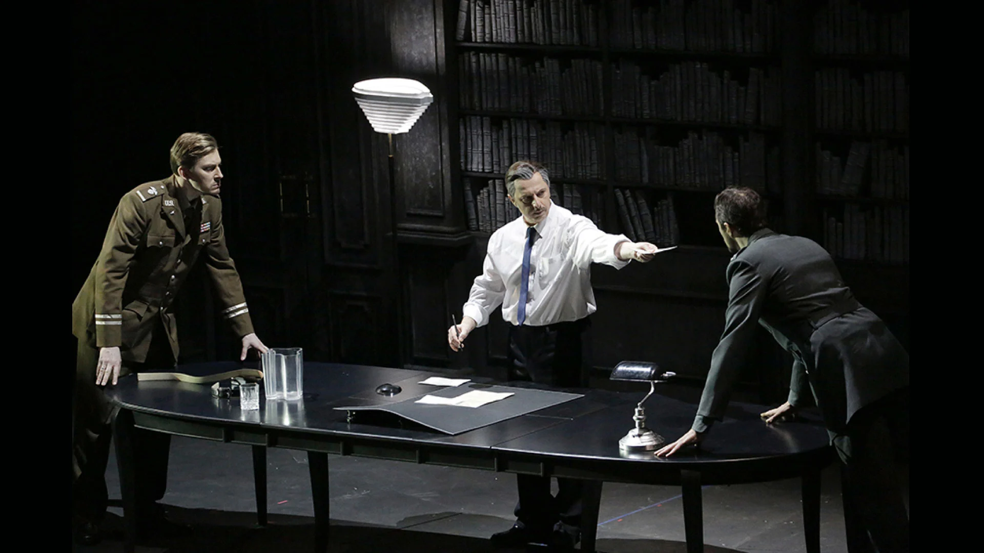 Three performers around a dark oval table in a study lined with bookshelves, lit by a standard lamp – Un Ballo in Maschera at the Bolshoi Theatre, Moscow, Russia, 2018 – opera set design