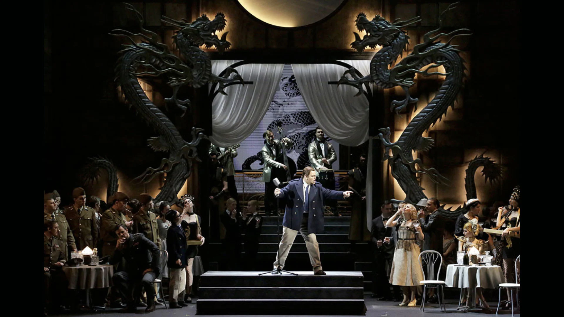 Stage set with two giant dragon sculptures framing a draped doorway, soldiers and guests at café tables – Un Ballo in Maschera at the Bolshoi Theatre, Moscow, Russia, 2018 – opera set design