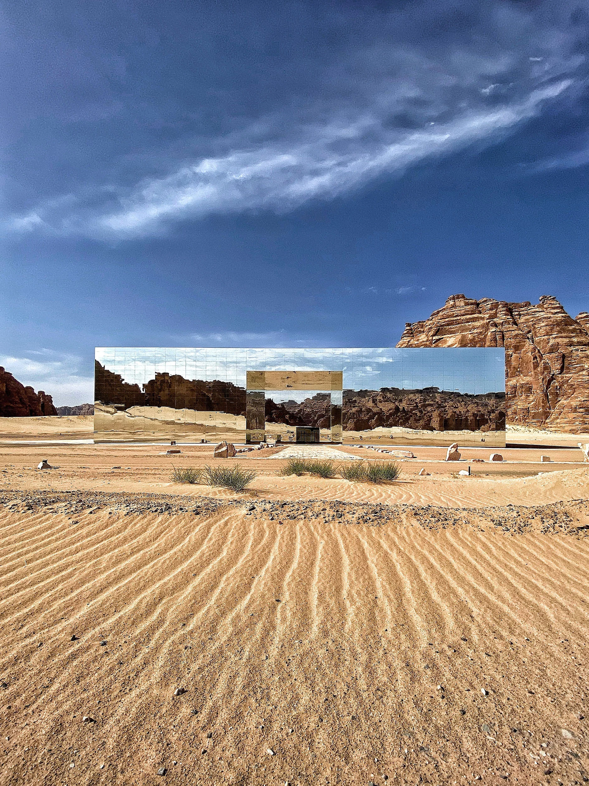 Mirrored facade seen across rippled sand dunes, reflecting the desert and rocks beneath a blue sky – Maraya Concert Hall, AlUla, Saudi Arabia, 2019 – architecture