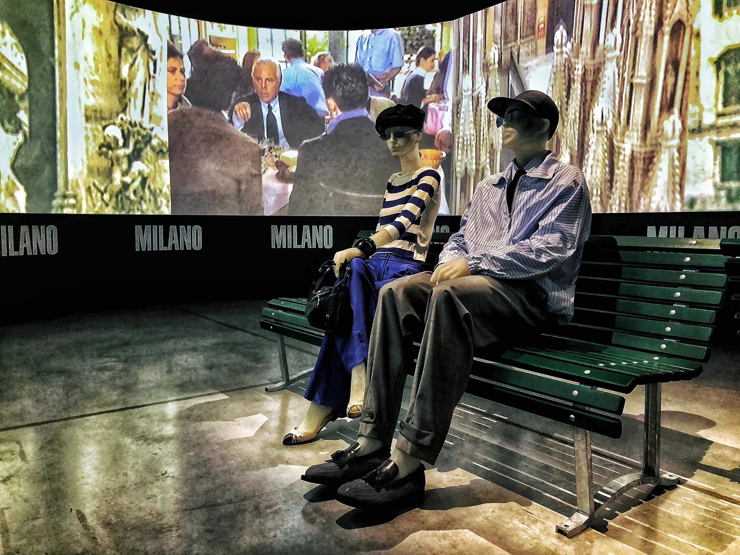 Two mannequins on a green park bench before a curved screen with Milan scenes and Milano lettering – Emporio Armani The Way We Are, Milan, Italy, 2020 – exhibition design