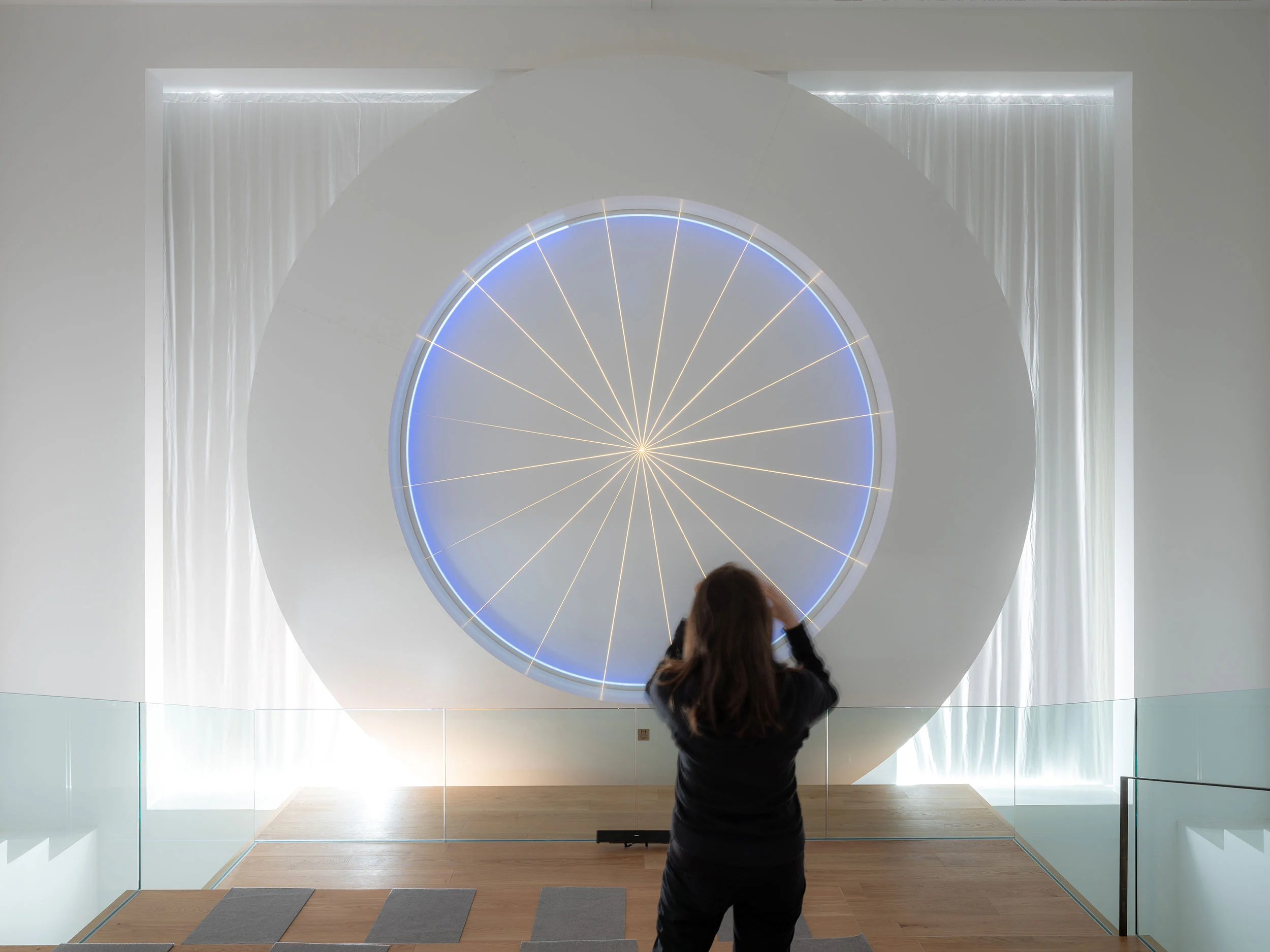 Visitor photographing a circular aperture lit in blue with radiating lines, set in a white wall between curtains – Lumen Museum of Mountain Photography, Kronplatz, Italy, 2018 – architecture
