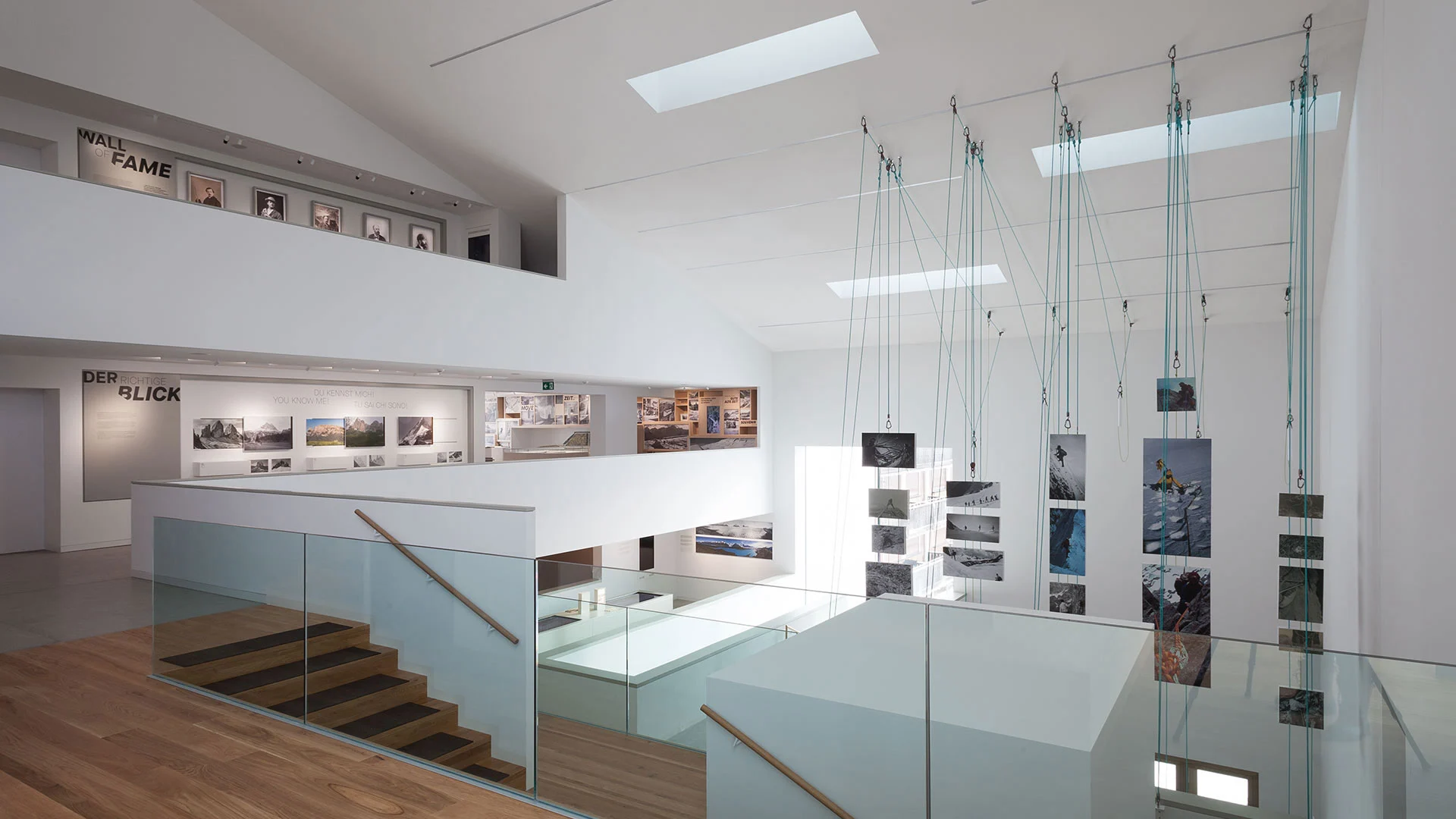 Bright exhibition space with photographs suspended on cables over a glass-railed stair and galleries along the walls – Lumen Museum of Mountain Photography, Kronplatz, Italy, 2018 – architecture