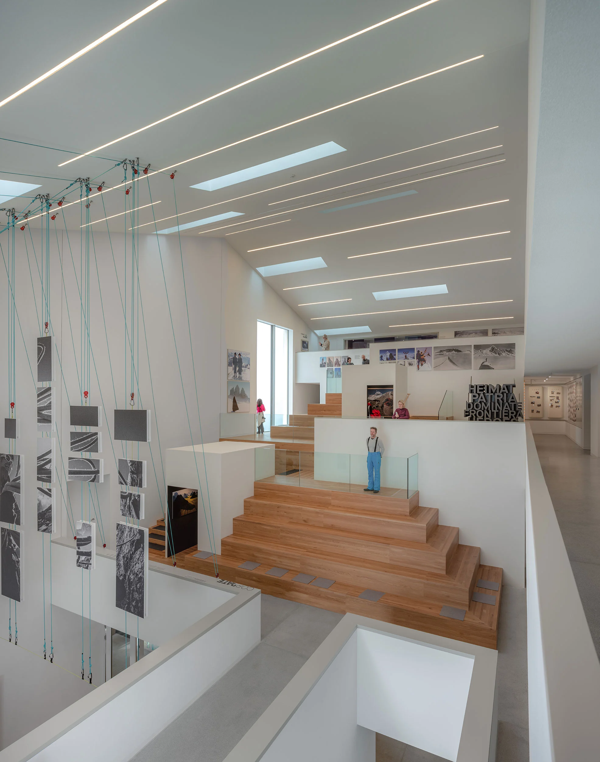 Multi-level museum interior with wooden stepped seating, linear ceiling lights and photographs hung on cables – Lumen Museum of Mountain Photography, Kronplatz, Italy, 2018 – architecture