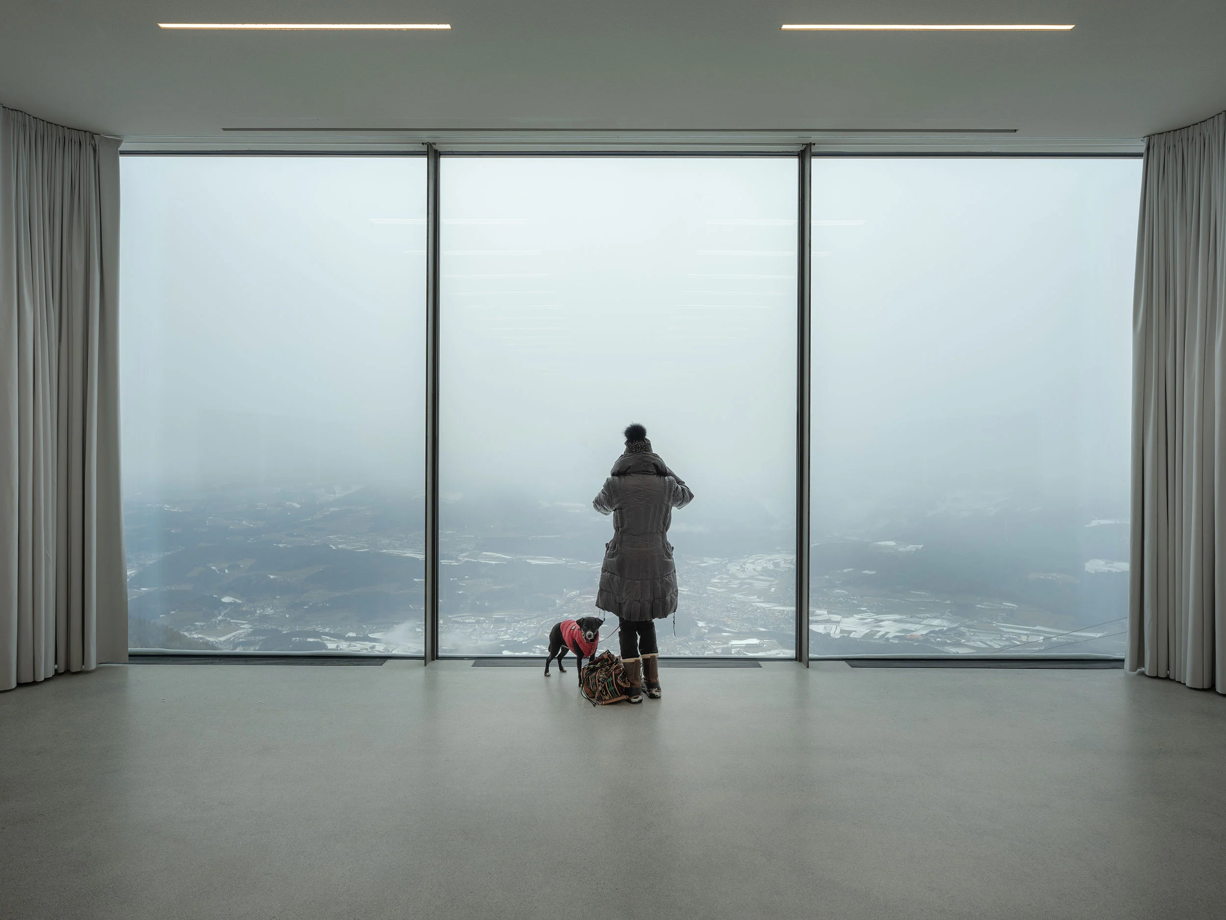 Visitor with a dog at a full-height window looking out over a misty, snowy mountain landscape – Lumen Museum of Mountain Photography, Kronplatz, Italy, 2018 – architecture