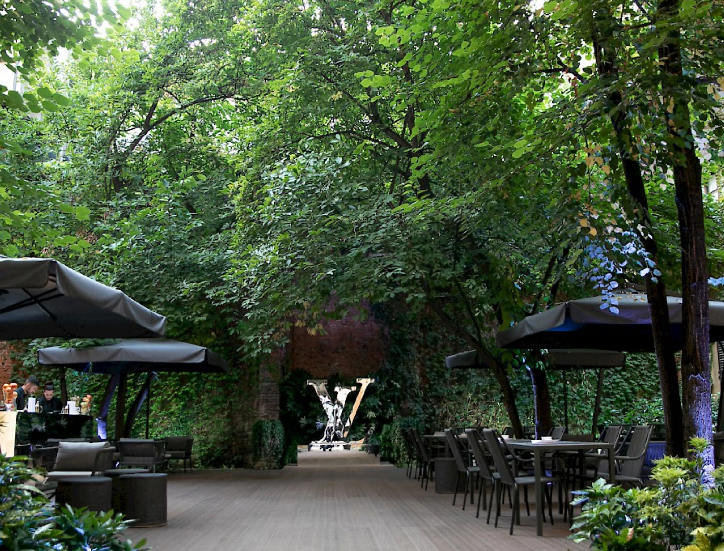 Garden lounge under tall trees with grey umbrellas, outdoor tables and an LV sculpture at the end – Louis Vuitton at Palazzo Clerici, Milan, Italy, 2019 – exhibition design
