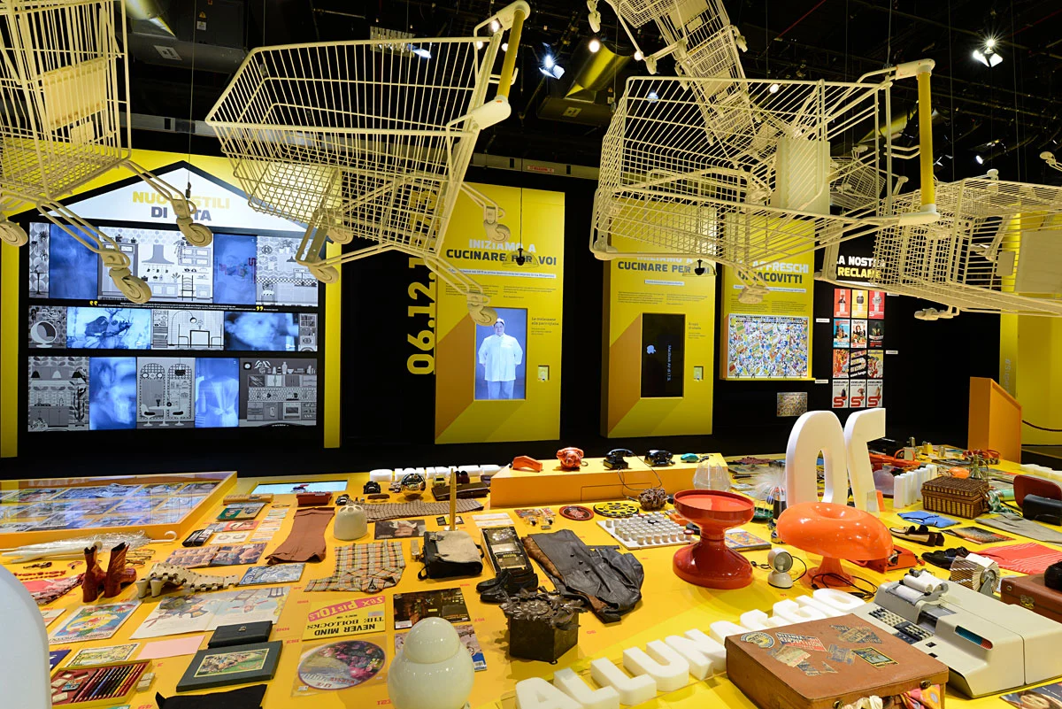 Supermostra Esselunga room with shopping trolleys hanging from the ceiling over a yellow table of vintage products – Esselunga Supermostra 60th Anniversary, Milan, Italy, 2018 – exhibition design
