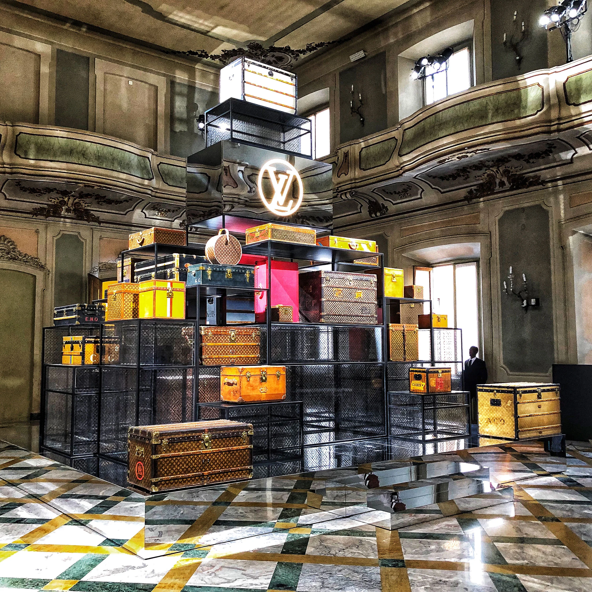 Pyramid of Louis Vuitton trunks on black mesh shelves under a lit LV logo, in a frescoed hall – Louis Vuitton at Palazzo Clerici, Milan, Italy, 2019 – exhibition design