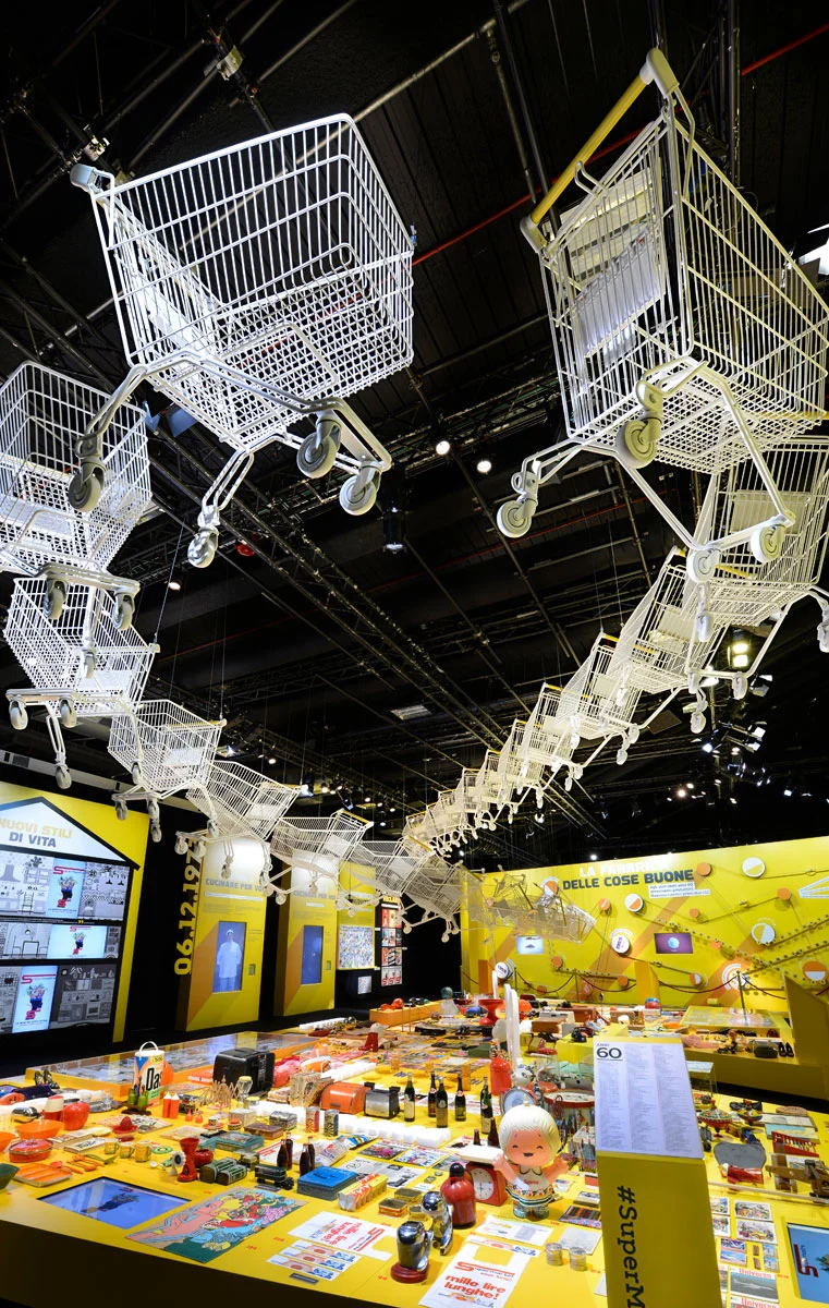 Vertical view of white shopping trolleys suspended in a long row above yellow display tables – Esselunga Supermostra 60th Anniversary, Milan, Italy, 2018 – exhibition design