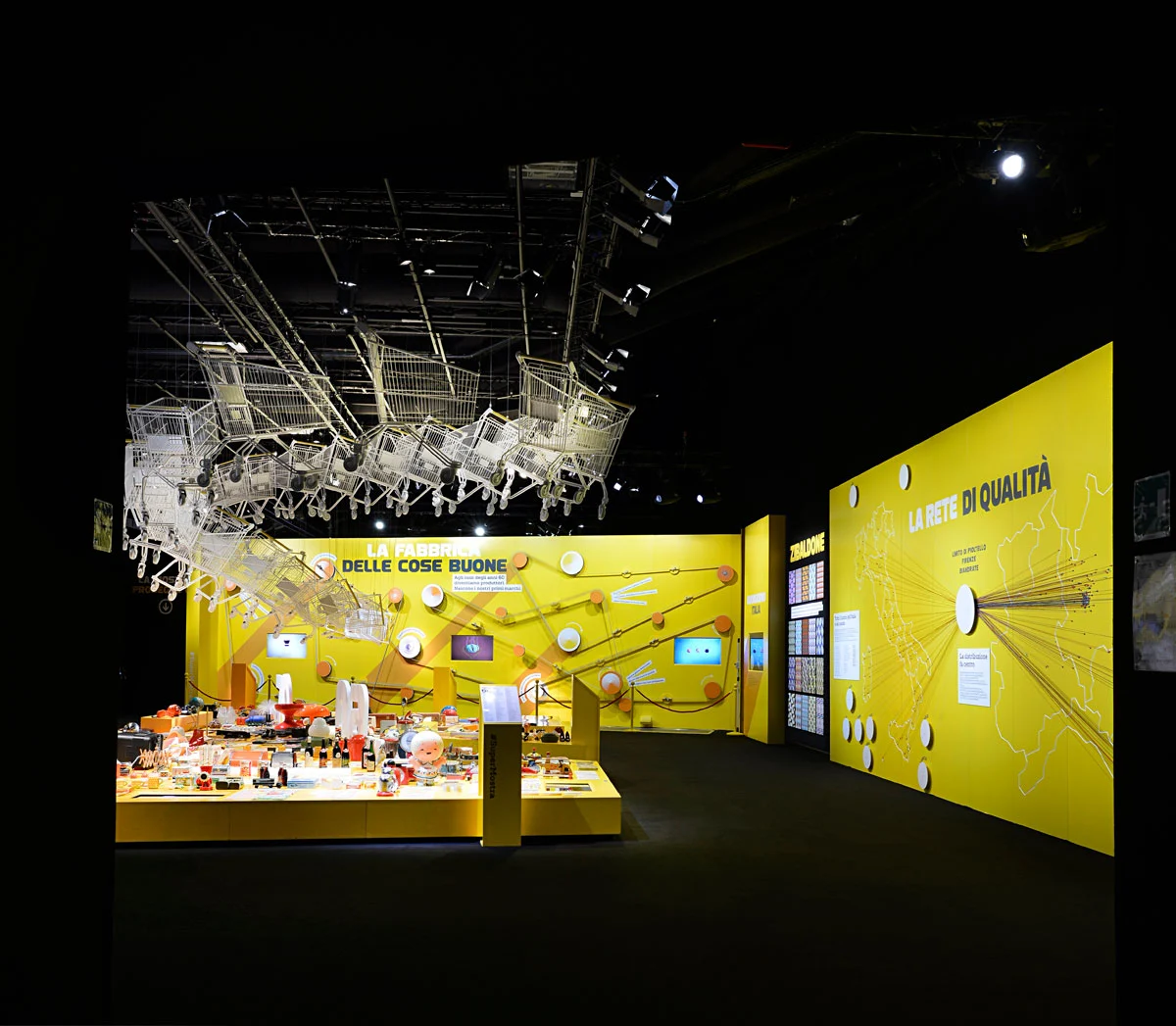 Dark exhibition hall with a cloud of hanging trolleys over a yellow display and a wall map titled La rete di qualità – Esselunga Supermostra 60th Anniversary, Milan, Italy, 2018 – exhibition design
