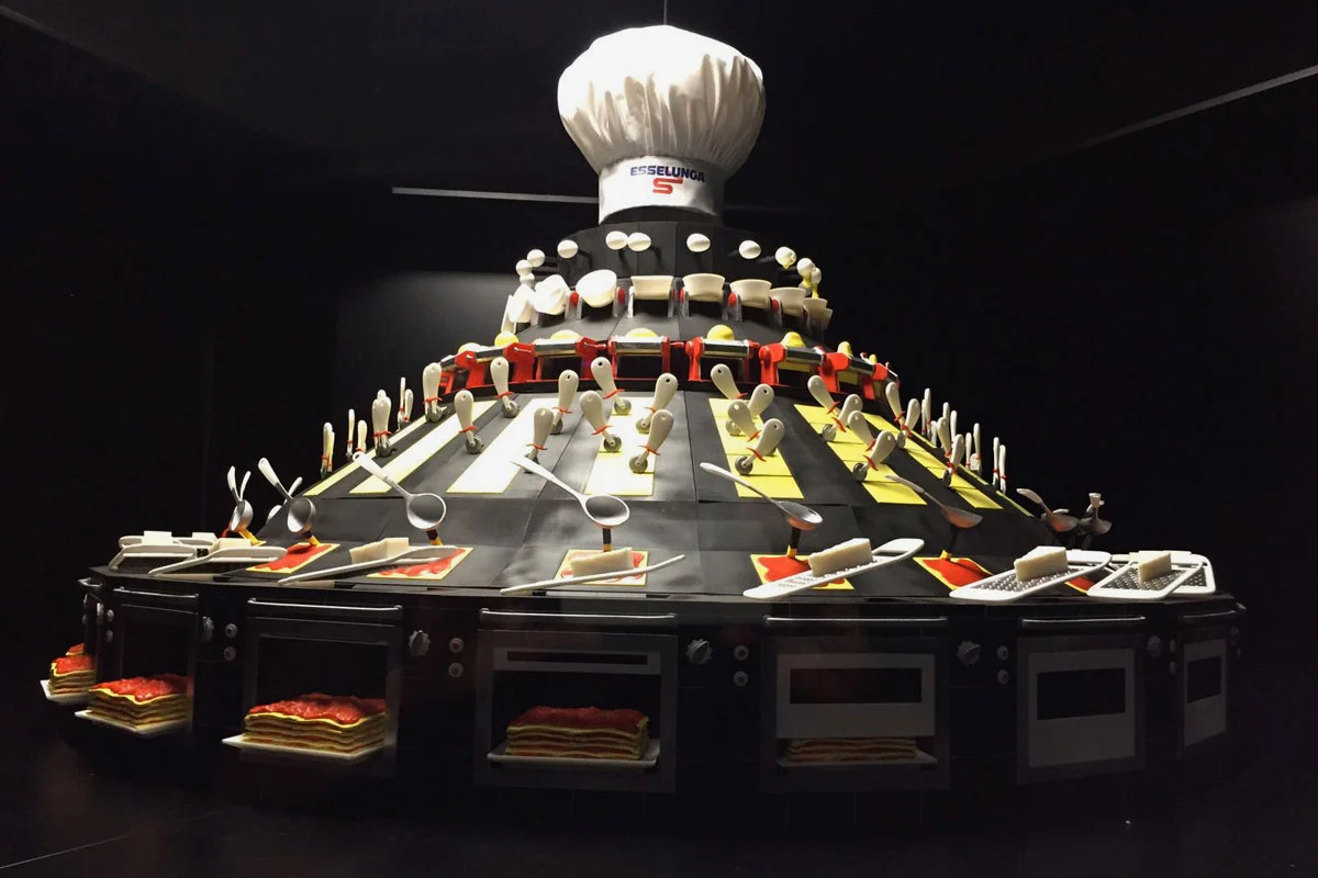 Tiered sculpture topped by a giant Esselunga chef's hat, with utensils, trays of lasagna and oven doors – Esselunga Supermostra 60th Anniversary, Milan, Italy, 2018 – exhibition design