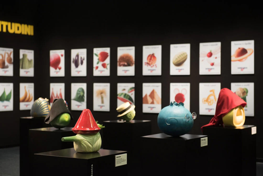 Vegetable-shaped sculptures in hats on black plinths, in front of a wall of illustrated posters – Esselunga Supermostra 60th Anniversary, Milan, Italy, 2018 – exhibition design