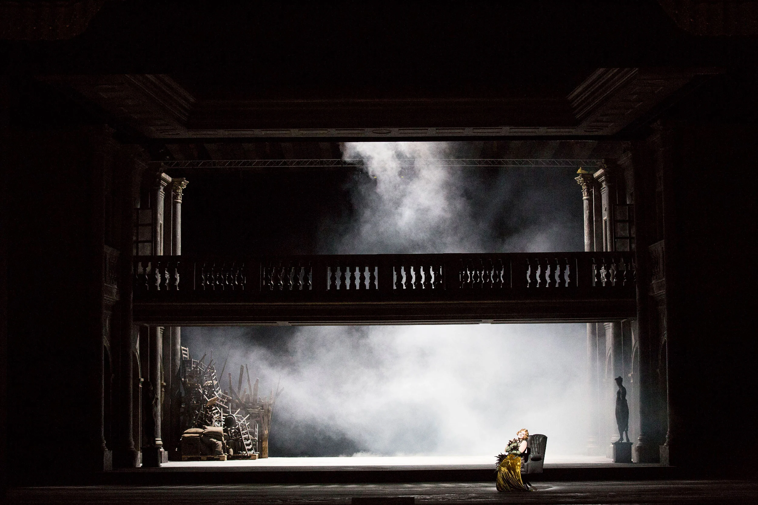 Dark stage with a balustraded gallery and smoke, a lone figure seated in an armchair in a pool of light – Tamerlano at La Scala, Milan, Italy, 2017 – opera set design
