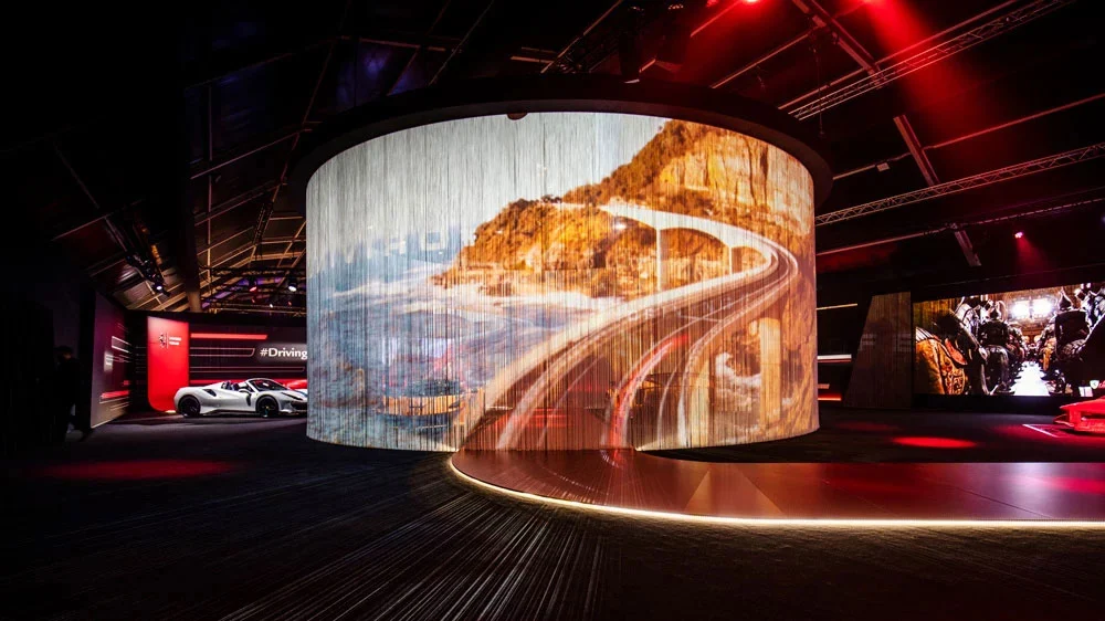 Cylindrical string-curtain screen showing a coastal road, ringed by light in a dark exhibition hall – Universo Ferrari at Fabbrica del Vapore, Milan, Italy, 2019 – exhibition design