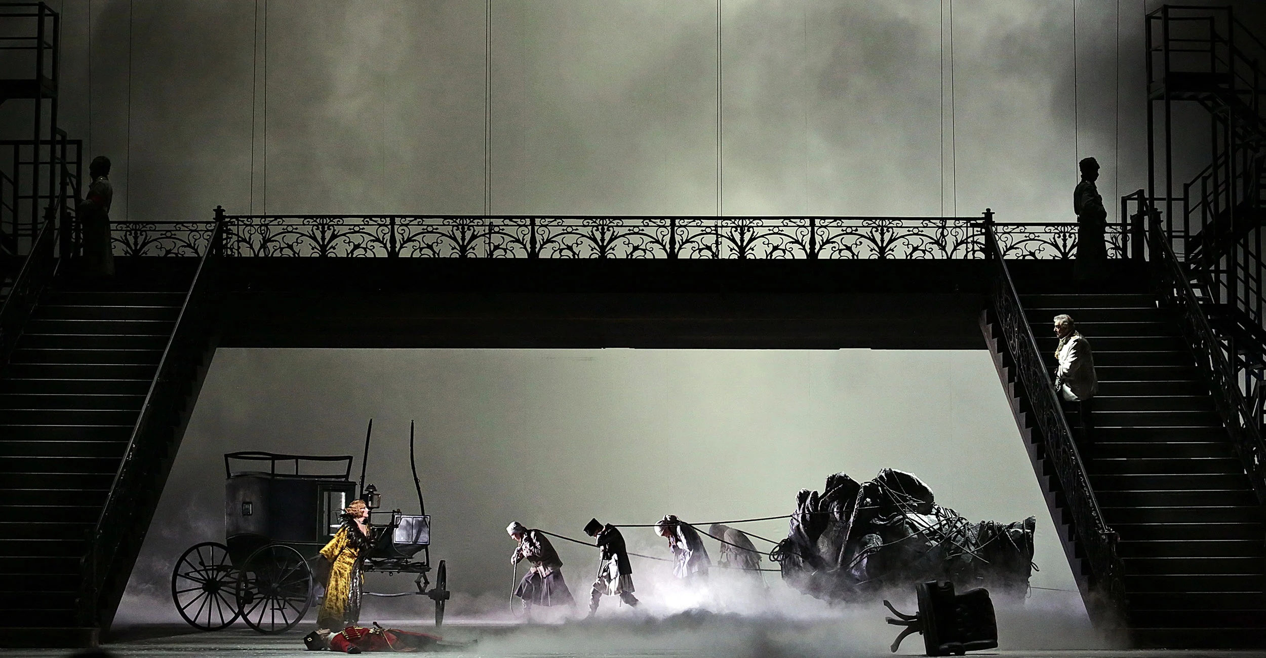 Iron footbridge with staircases over a misty stage, figures hauling a carriage and a fallen horse – Tamerlano at La Scala, Milan, Italy, 2017 – opera set design