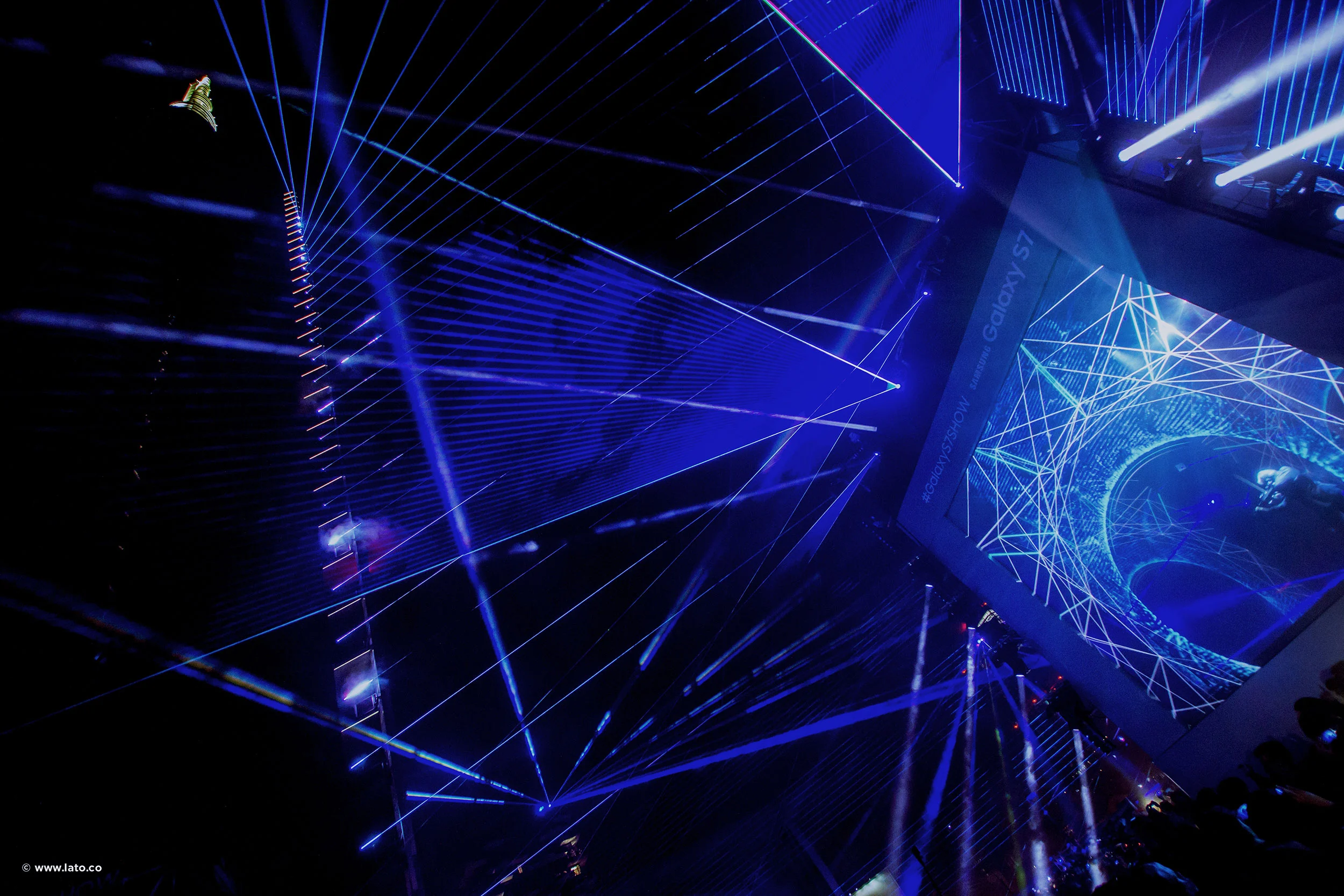 Upward view of blue laser lines forming geometric patterns between the buildings, beside a video screen – Samsung Galaxy S7 Laser Show, Milan, Italy, 2016 – event design
