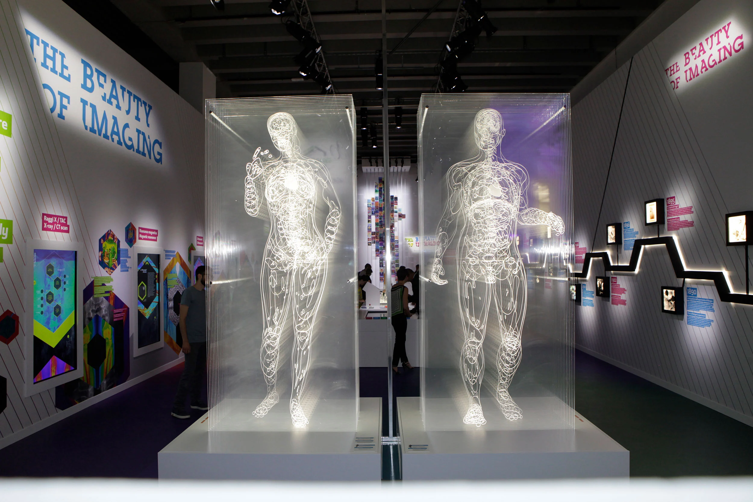 Front view of two glowing transparent anatomical figures, with colourful panels and a timeline along the walls – Bracco The Beauty of Imaging, Milan, Italy, 2017 – exhibition design