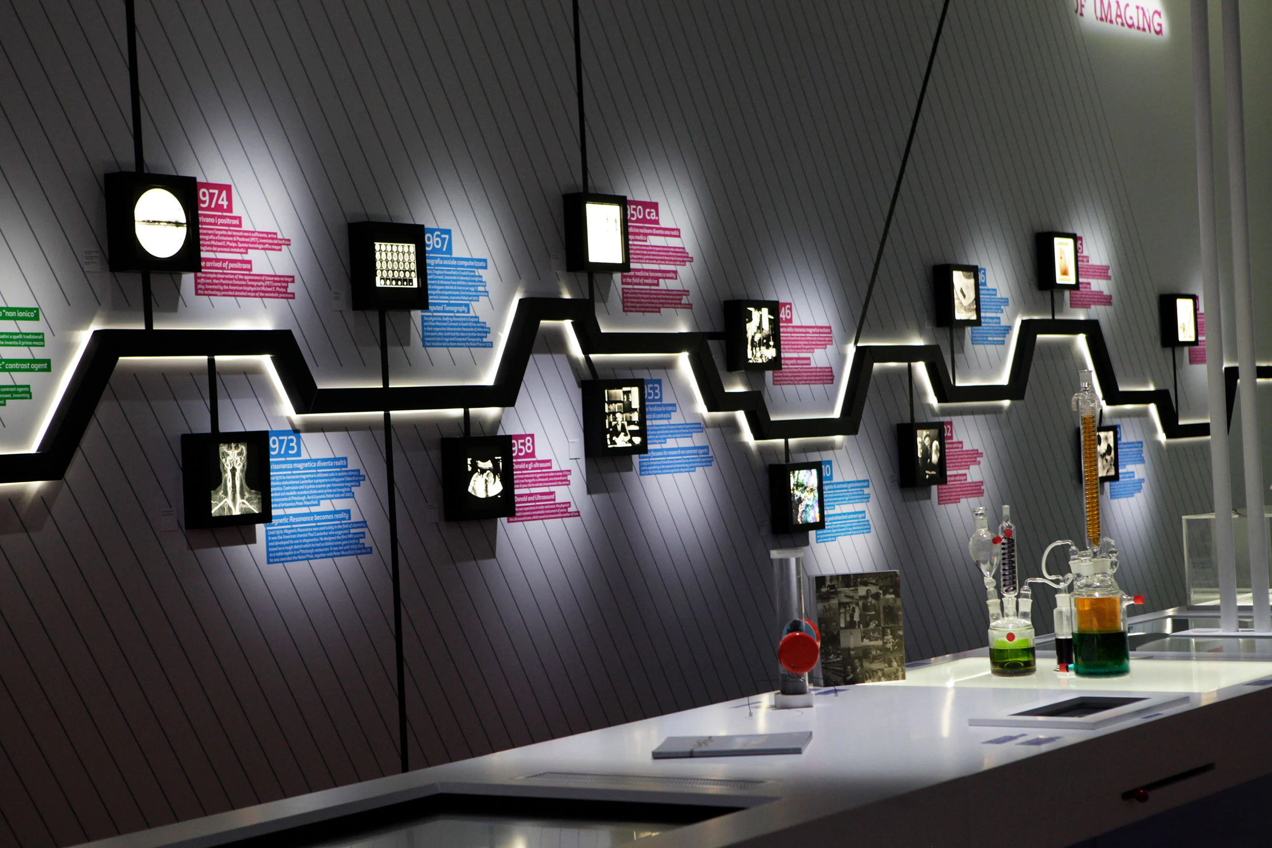 Wall timeline of backlit diagnostic images on a zigzag light line, with lab glassware on the table below – Bracco The Beauty of Imaging, Milan, Italy, 2017 – exhibition design