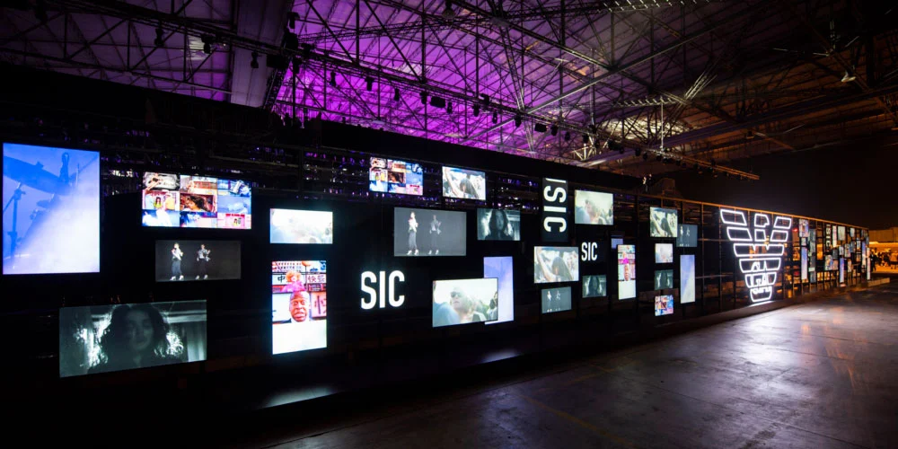 Long black wall of video screens with SIC signs and the eagle logo under purple-lit roof trusses – Emporio Armani Boarding SS19, Milan, Italy, 2019 – fashion show set design