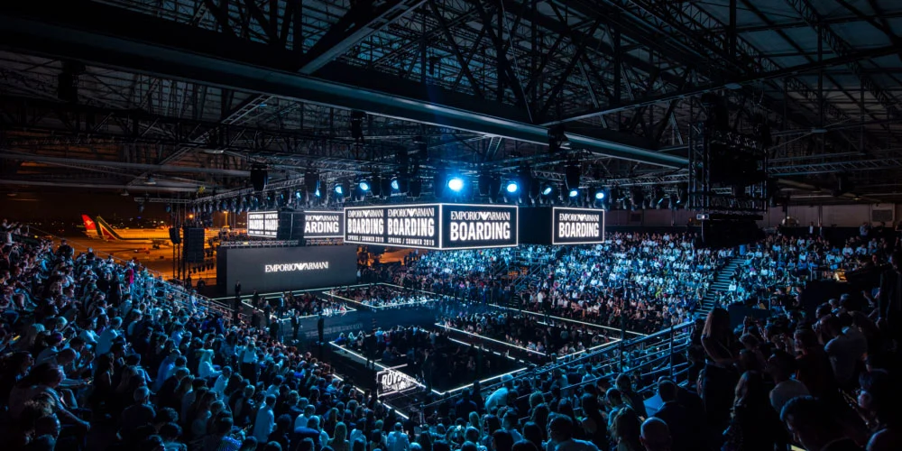 Wide view of the hangar with tiered seating, Emporio Armani Boarding screens and a plane in the background – Emporio Armani Boarding SS19, Milan, Italy, 2019 – fashion show set design