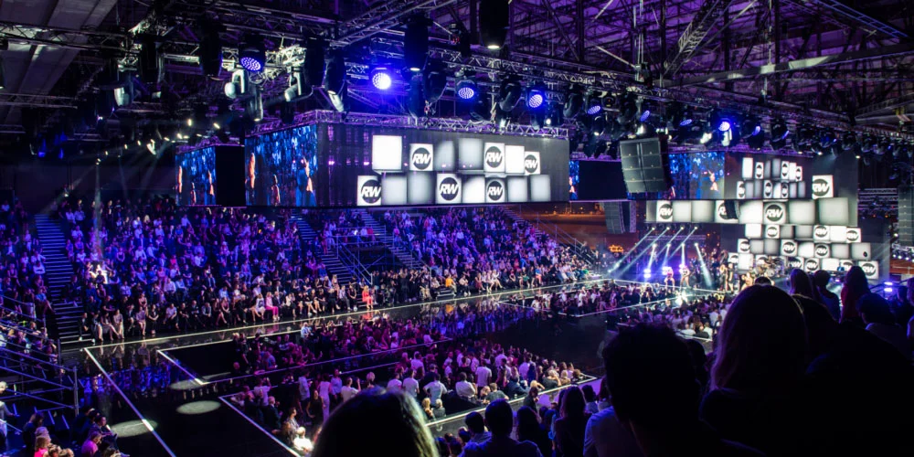 Hangar packed with spectators in blue light, runways and suspended screens showing RW logos – Emporio Armani Boarding SS19, Milan, Italy, 2019 – fashion show set design