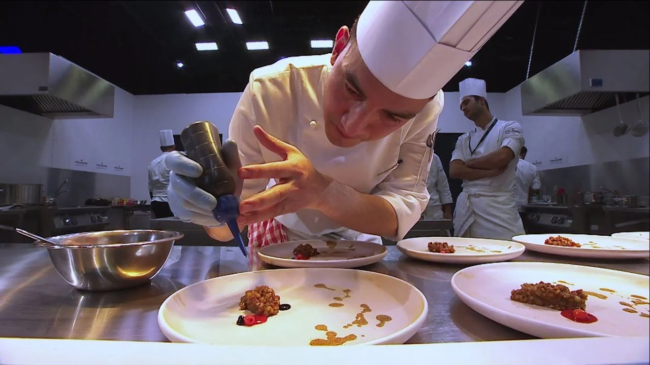 Young chef in a white hat plating dishes with a squeeze bottle in a professional kitchen – S.Pellegrino Young Chef, Milan, Italy, 2019 – event design