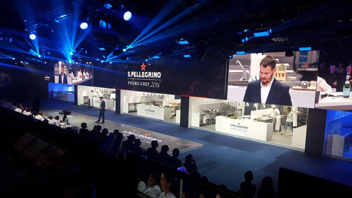 Stage of open kitchen stations with a large screen showing a close-up and blue spotlights – S.Pellegrino Young Chef, Milan, Italy, 2019 – event design