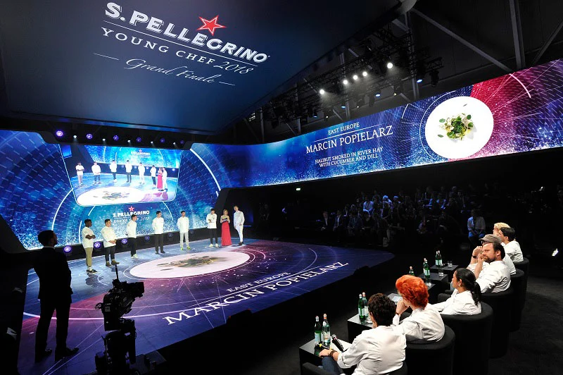 Grand final stage with a curved LED wall showing a dish and finalist names, jury seated at right – S.Pellegrino Young Chef, Milan, Italy, 2019 – event design