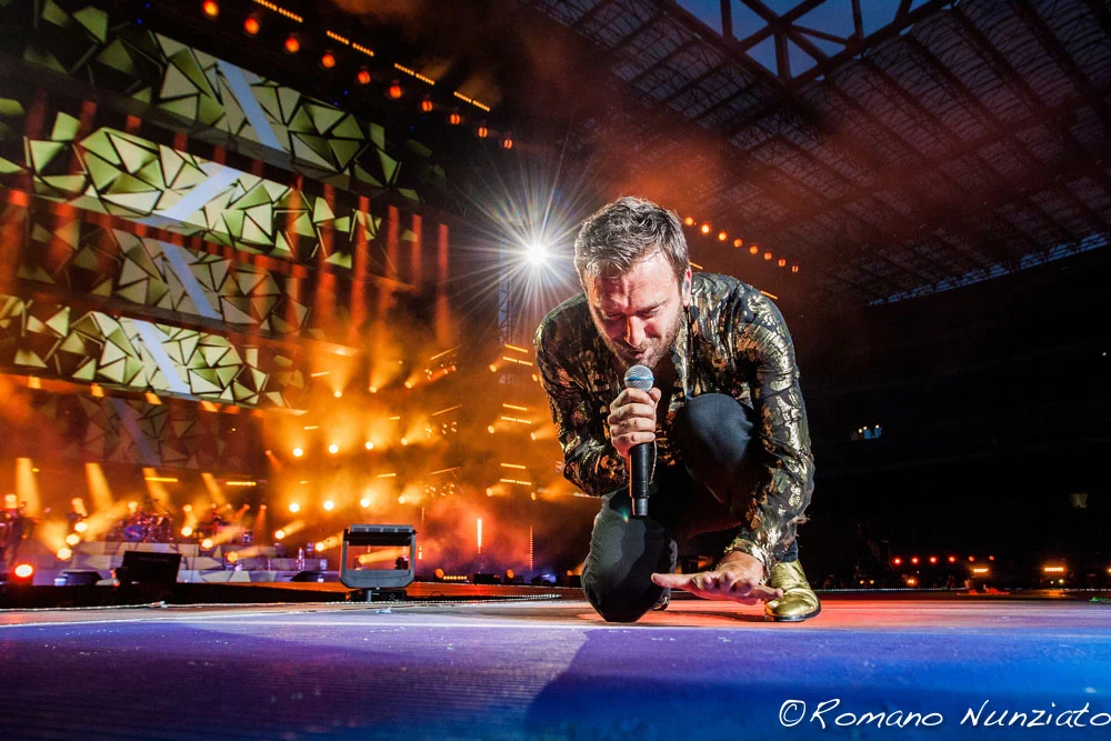 Cesare Cremonini kneeling with a microphone, geometric video screens in orange light behind – Cesare Cremonini Stadi 2018, Italy – concert stage design
