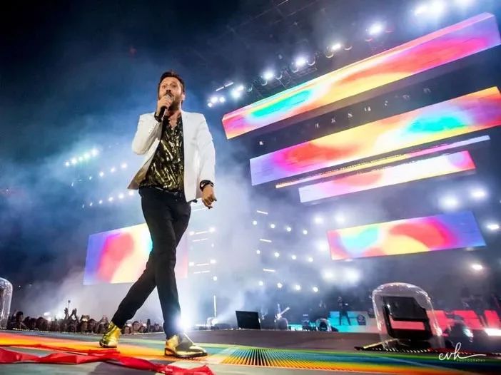Cesare Cremonini singing at the stage edge in a white jacket, colourful video bars behind him – Cesare Cremonini Stadi 2018, Italy – concert stage design