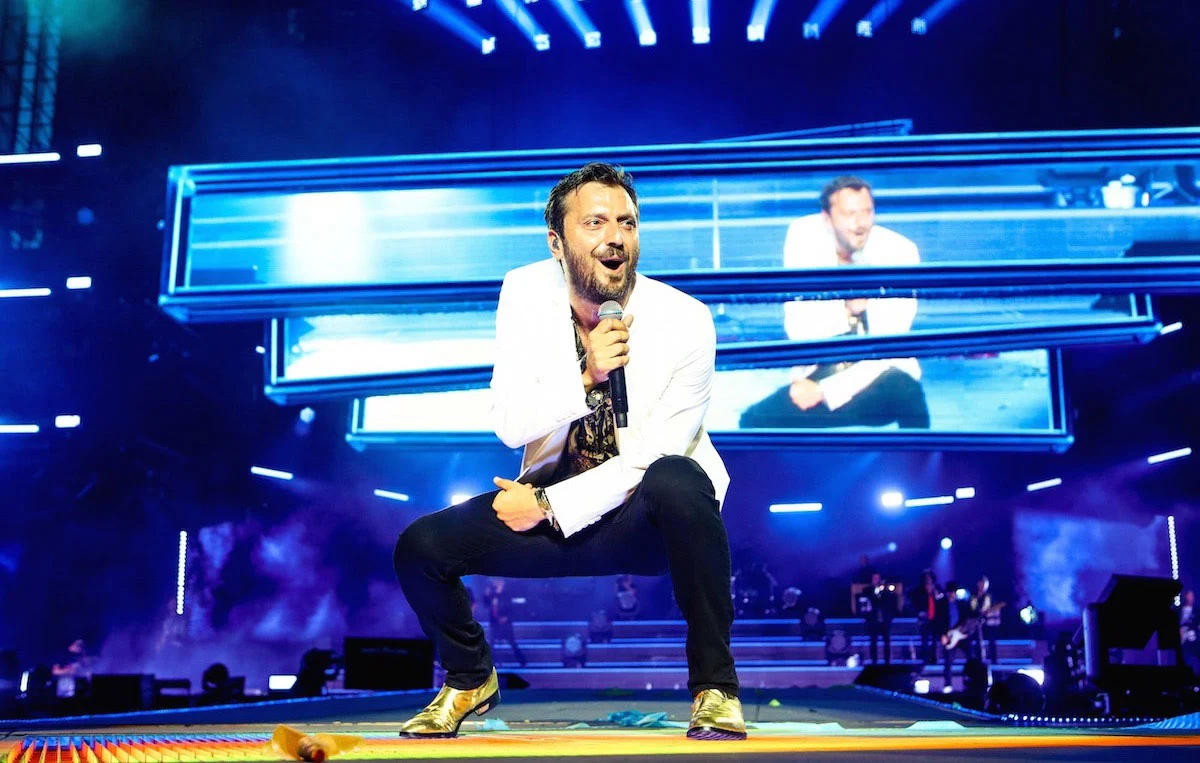Cesare Cremonini crouching on stage, his image on a stack of blue video bars behind him – Cesare Cremonini Stadi 2018, Italy – concert stage design