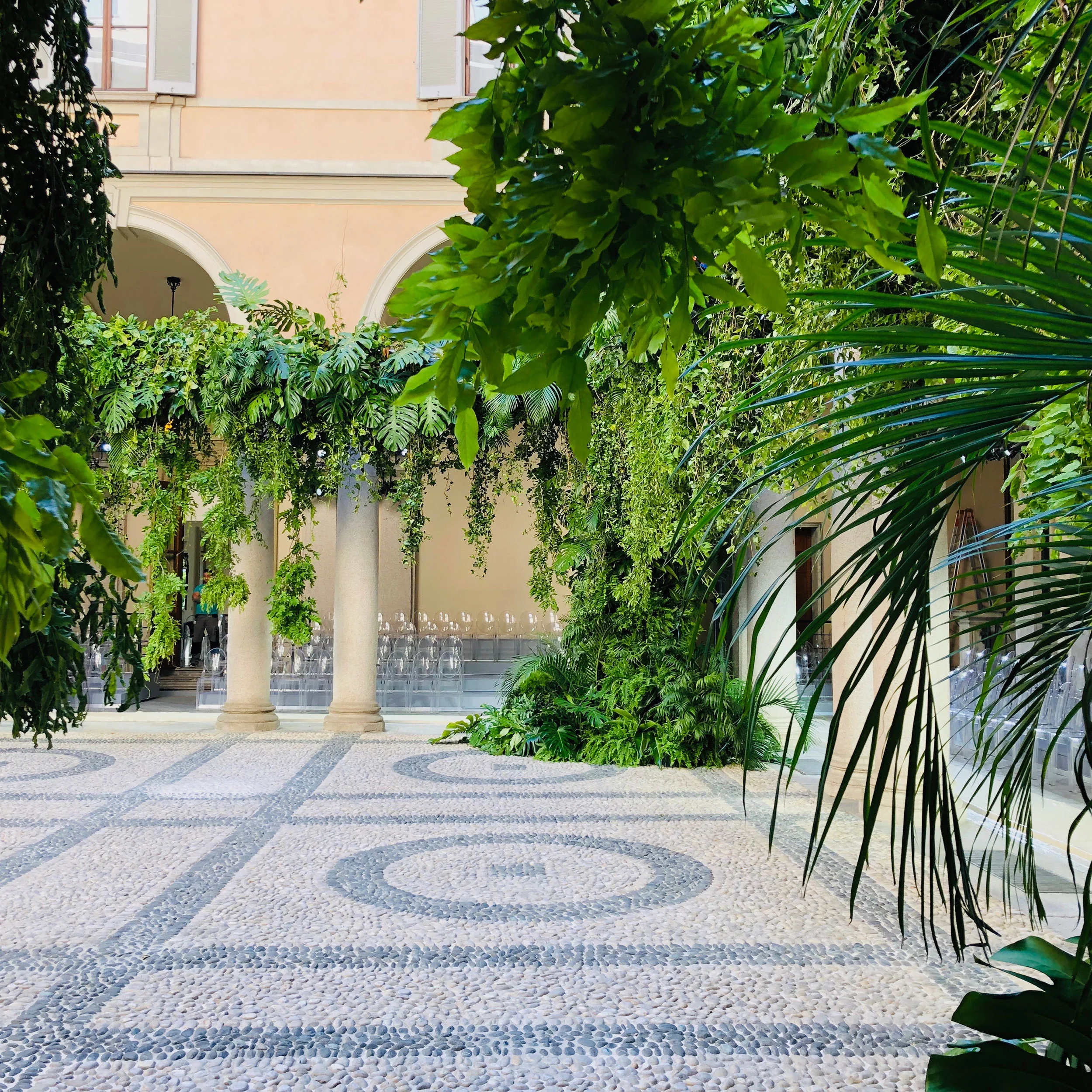 Palazzo Orsini cloister with cobbled floor, columns covered in tropical plants and rows of clear chairs – Giorgio Armani SS20 Men and Women, Milan, Italy, 2019 – fashion show set design