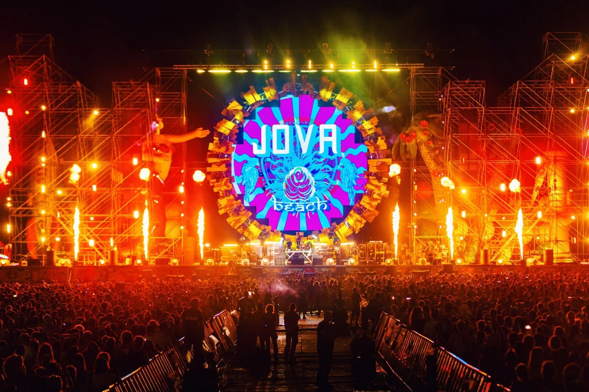 Night view of the Jova Beach stage: circular logo screen between scaffold towers, flames and a big crowd – Jova Beach Party 2019, Italy – concert stage design