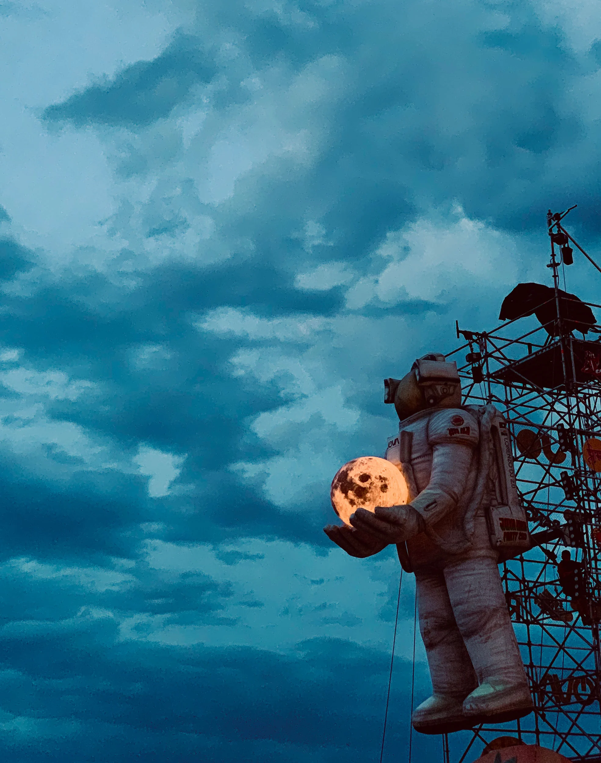 Inflatable astronaut holding a glowing moon on the scaffold tower against a cloudy evening sky – Jova Beach Party 2019, Italy – concert stage design
