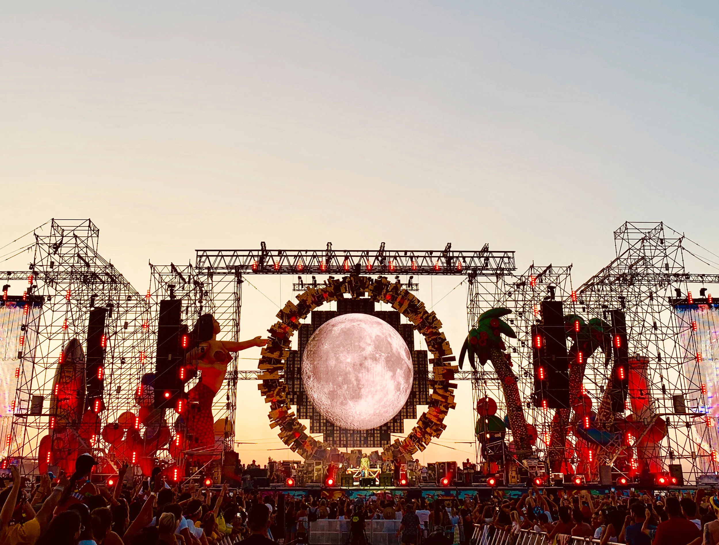 Stage at dusk with a full moon image inside the ring, mermaid and palm tree figures on red-lit towers – Jova Beach Party 2019, Italy – concert stage design