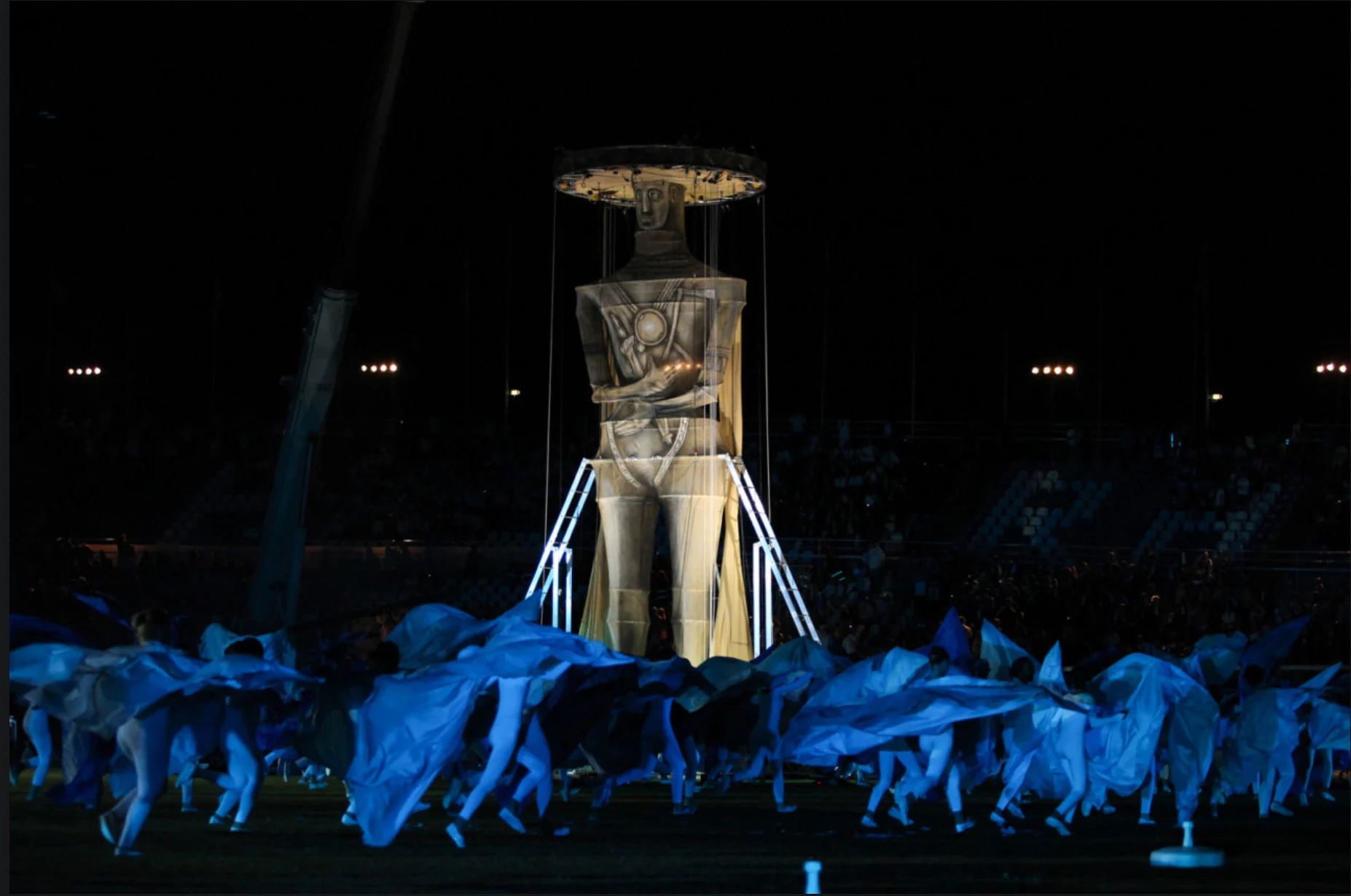 Giant illuminated figure on a frame above performers waving blue fabric in the stadium – Pescara 2009 Mediterranean Games Opening Ceremony, Italy, 2019 – event design