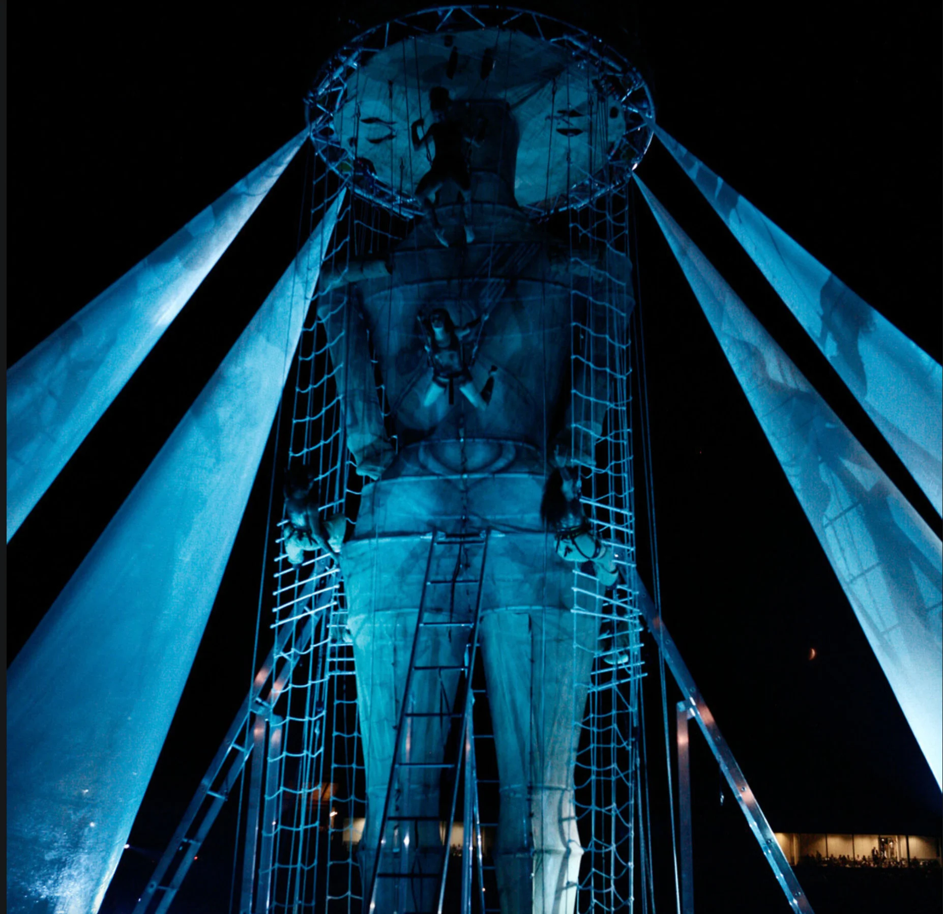 Upward view of the giant figure inside its net and ladder structure, flanked by blue-lit fabric sails – Pescara 2009 Mediterranean Games Opening Ceremony, Italy, 2019 – event design