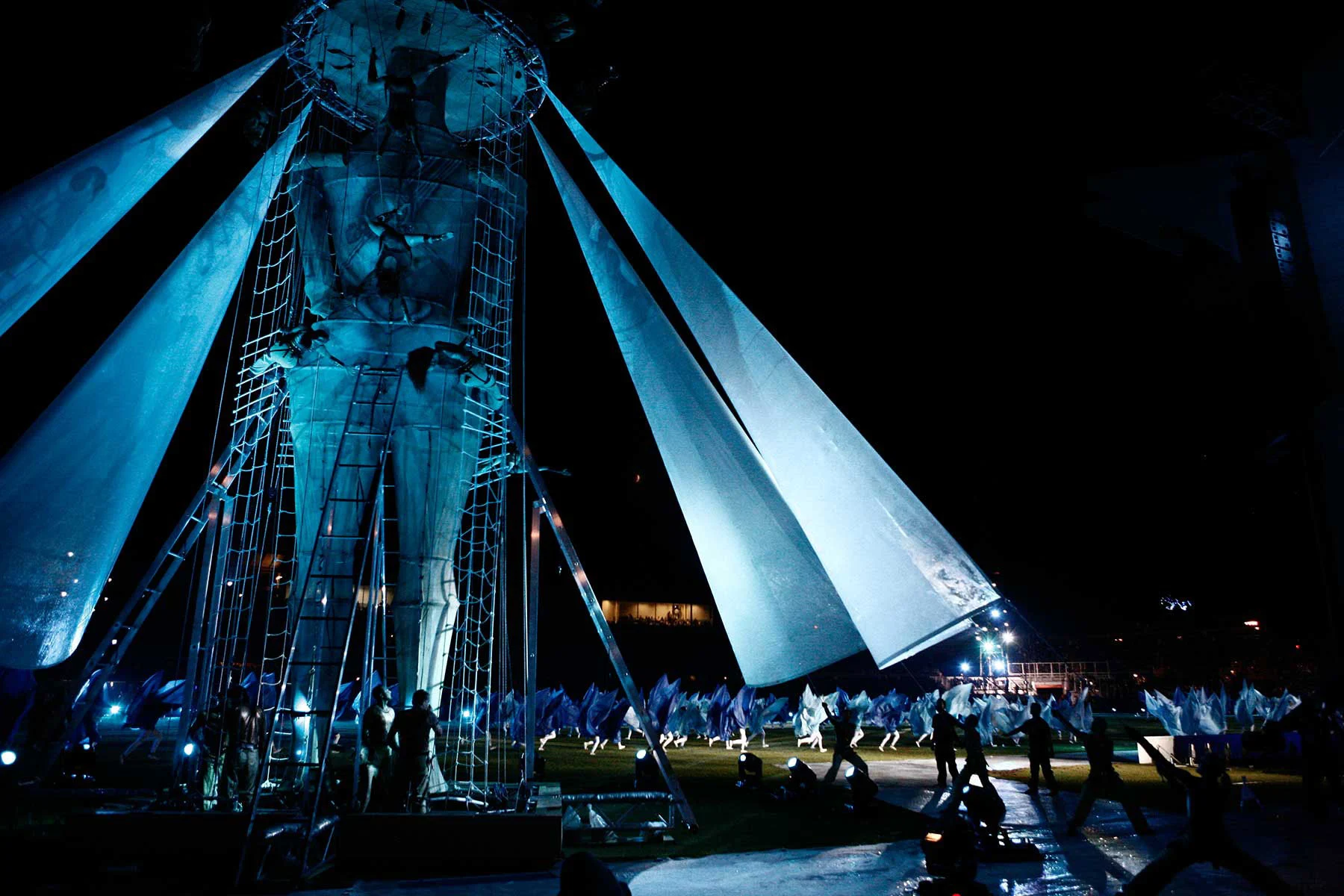 Tall statue-like figure in a scaffold of ladders and nets with sail-like fabric wings, lit blue – Pescara 2009 Mediterranean Games Opening Ceremony, Italy, 2019 – event design