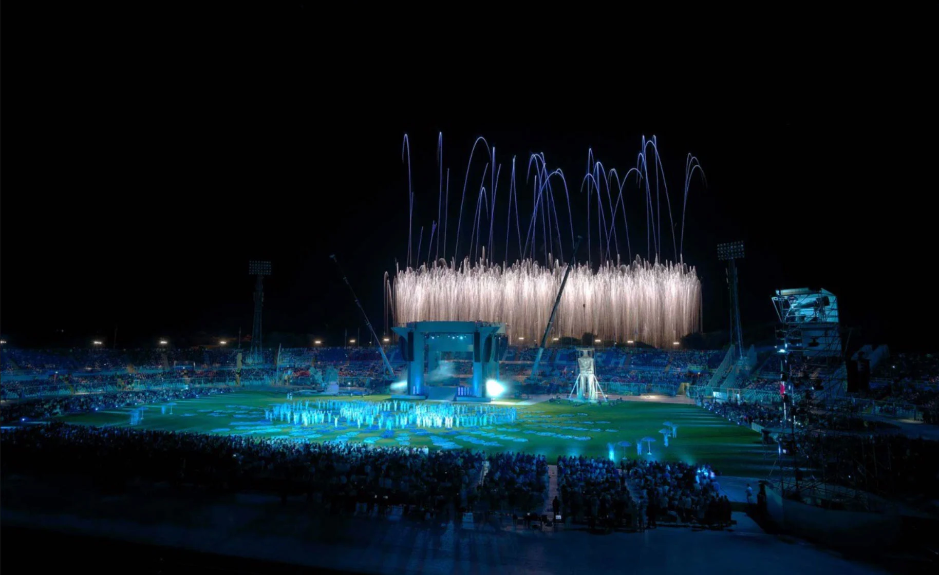 Night view of the stadium with fireworks fountains above a blue-lit field and the audience – Pescara 2009 Mediterranean Games Opening Ceremony, Italy, 2019 – event design