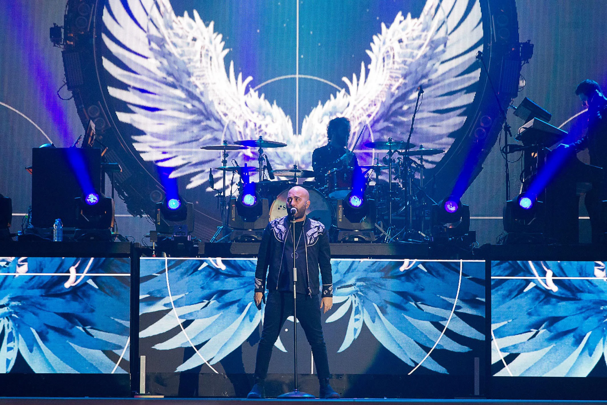 Negramaro singer at the front of the stage, white feathered wings projected on the screens behind the drum kit – Negramaro Amore che Torni Tour 2018, Italy – concert stage design
