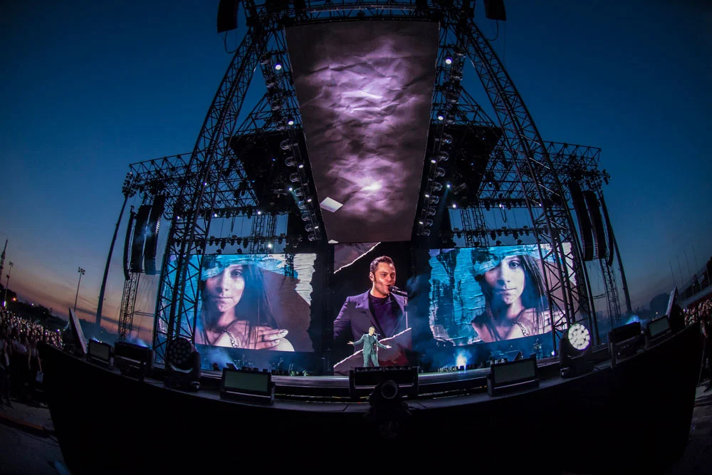Tiziano Ferro on stage at dusk under a steel tower, close-ups of the singer and a woman on the screens – Tiziano Ferro Il Mestiere della Vita Tour 2017, Italy – concert stage design