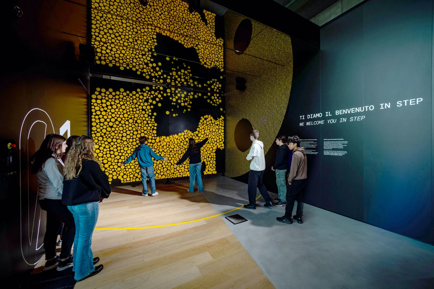 Visitors at an interactive wall of yellow dots responding to their silhouettes, beside a welcome text panel – STEP FuturAbility District by Fastweb, Milan, Italy, 2022 – exhibition design