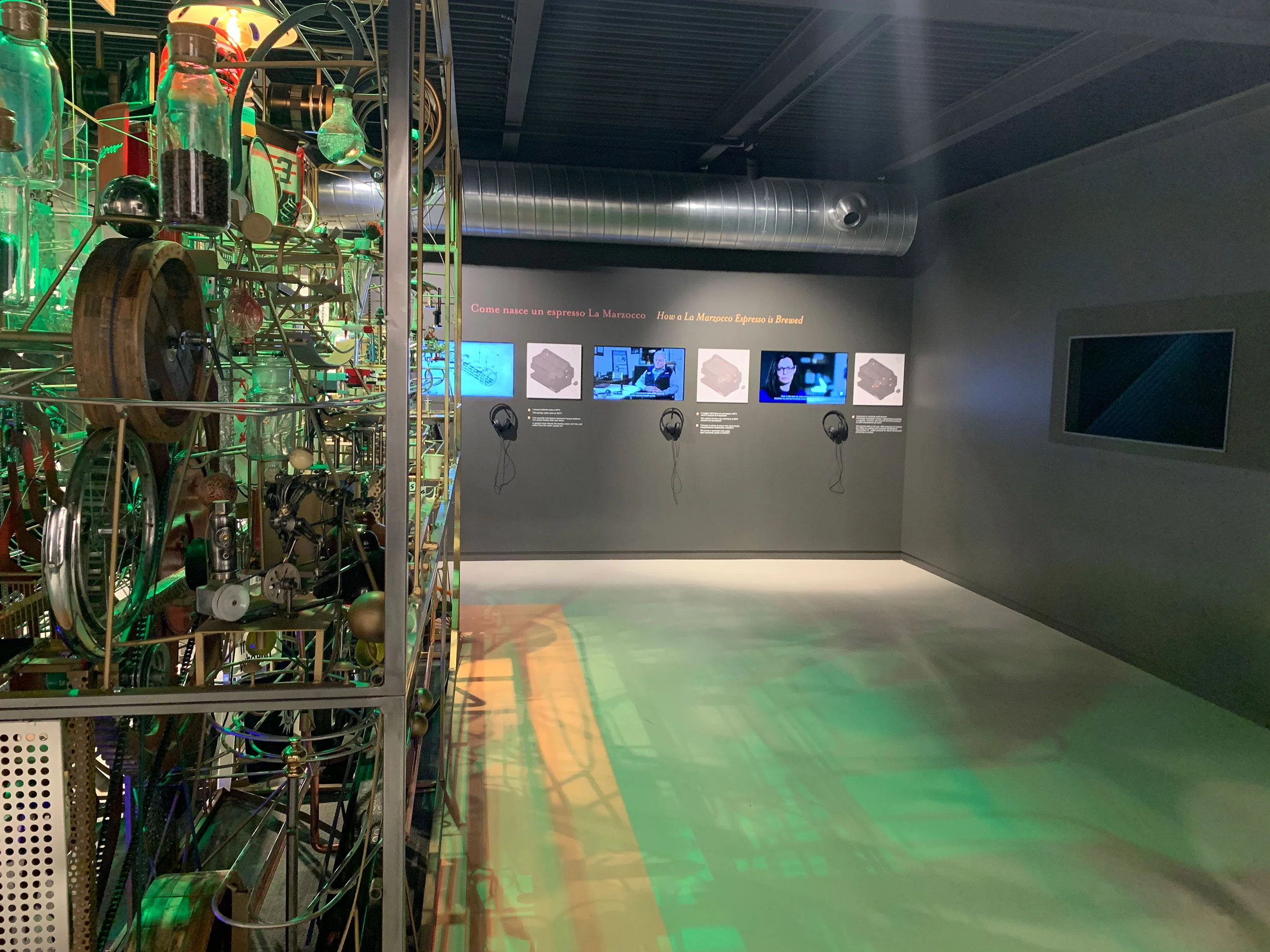 Dark exhibition room with a kinetic machine sculpture, video screens and headphones on the wall – La Marzocco Accademia del Caffè Espresso, Florence, Italy, 2018 – interior design