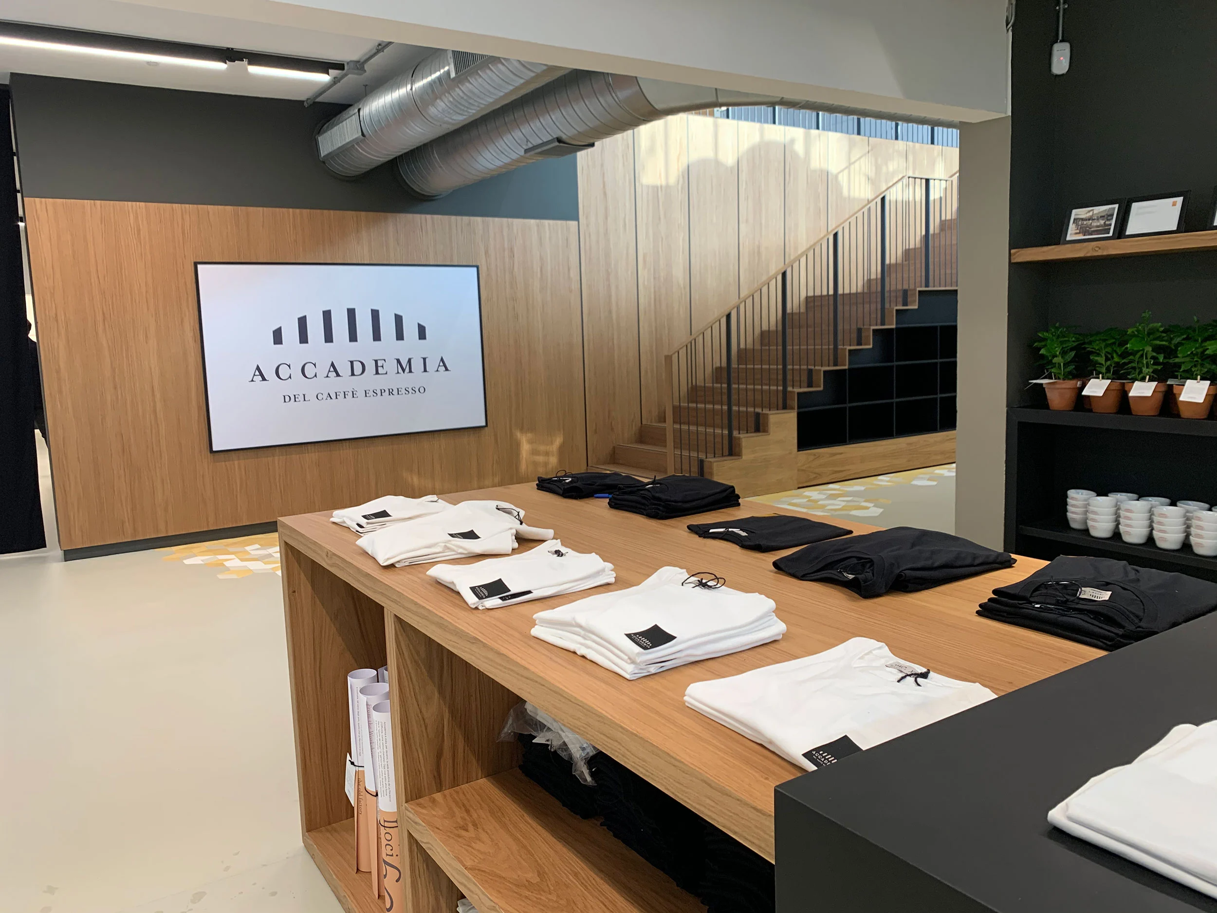 Shop with folded T-shirts on an oak table, a screen with the Accademia logo and a wooden staircase – La Marzocco Accademia del Caffè Espresso, Florence, Italy, 2018 – interior design
