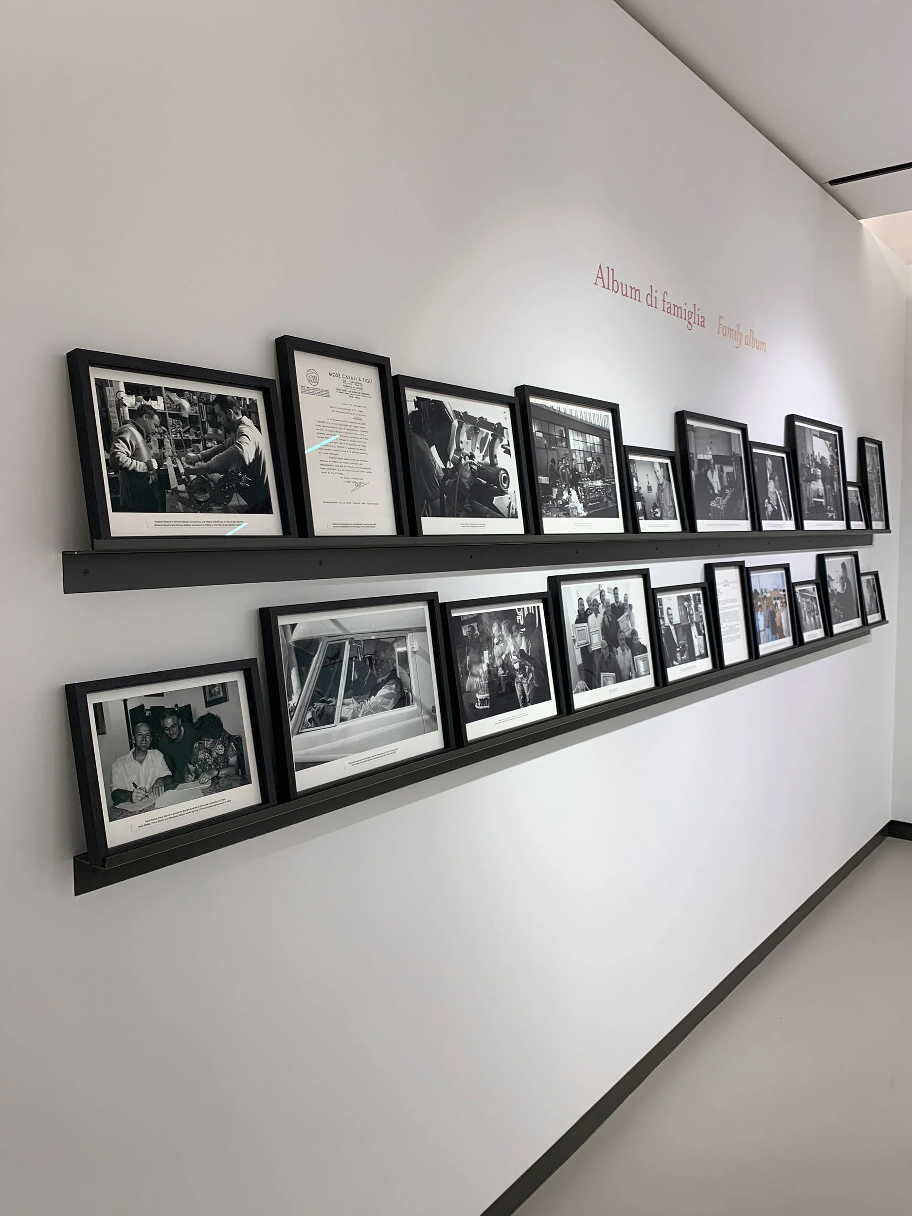 Corridor wall with two shelves of framed black and white photos under the title Album di famiglia – La Marzocco Accademia del Caffè Espresso, Florence, Italy, 2018 – interior design