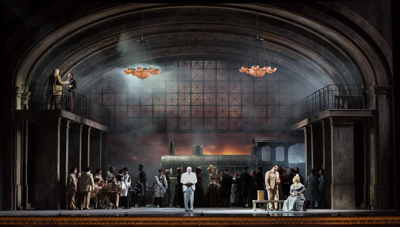 Railway station set with a vaulted iron-and-glass roof and a steam locomotive, chorus gathered on stage – Manon Lescaut at Teatro San Carlo, Naples, Italy, 2022 – opera set design